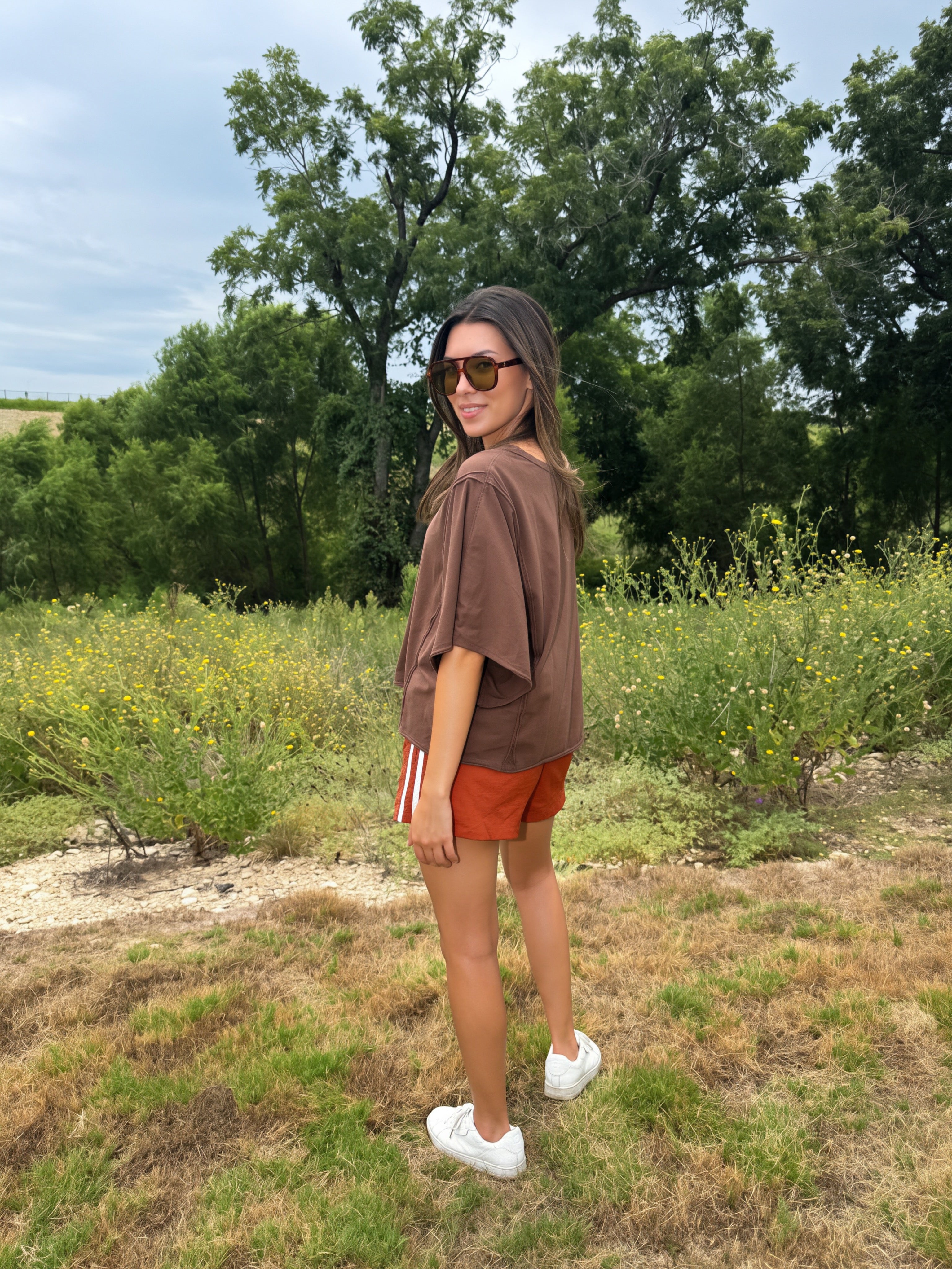 A woman wearing a brown oversized t-shirt, rust-orange shorts, white sneakers, fall fashion style , and stands outdoors in a grassy field with trees in the background.