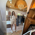A woman takes a mirror selfie in a boutique clothing store, wearing a beige and white striped matching lounge set consisting of a long-sleeve top and shorts, paired with white sneakers.