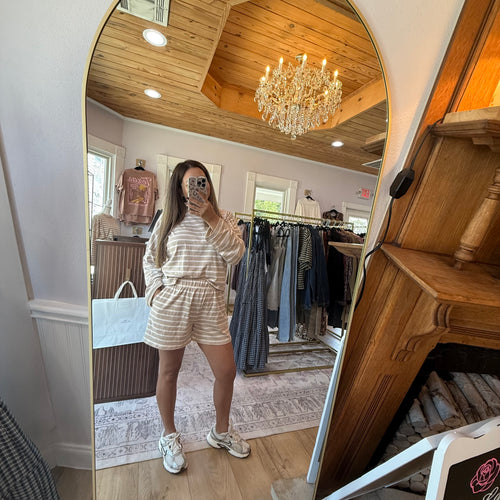 A woman takes a mirror selfie in a boutique clothing store, wearing a beige and white striped matching lounge set consisting of a long-sleeve top and shorts, paired with white sneakers.