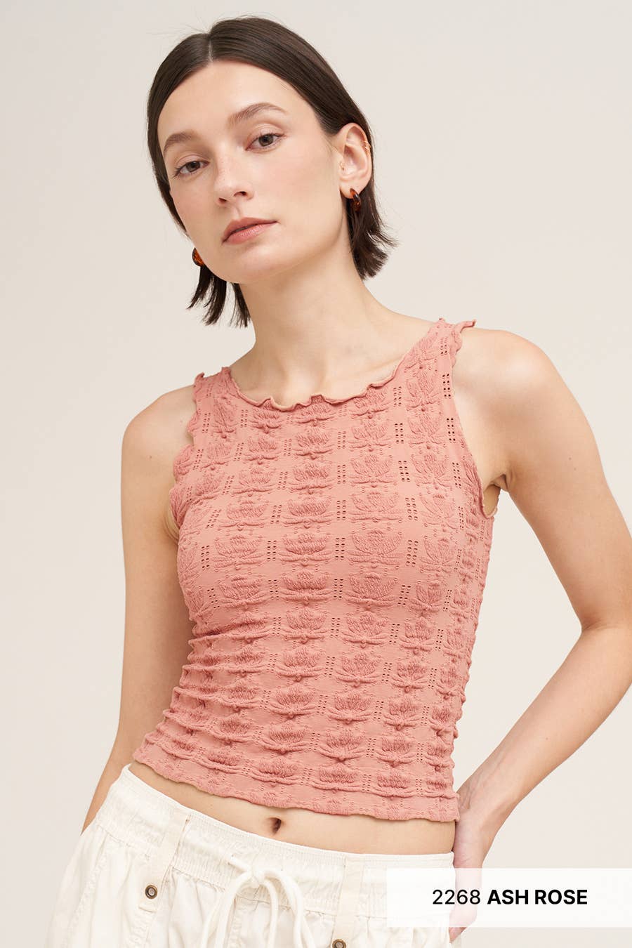 A woman wearing a textured, sleeveless cropped tank top in a dusty rose/ash rose color, paired with white drawstring pants, against a solid light beige background.