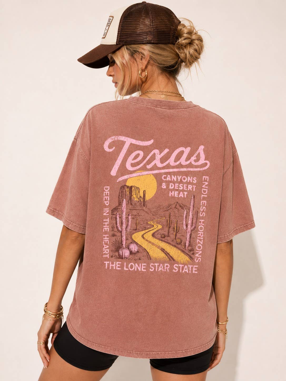 A woman shown from the back wearing a faded dusty rose oversized T-shirt with a large pink and yellow 'Texas' desert landscape graphic on the back, paired with a brown trucker hat and black shorts against a plain light beige background.
