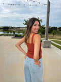 A woman wearing a burnt orange colored ribbed tank top and light blue jeans with gold star details, standing outdoors on a concrete patio with a lake and trees in the background.