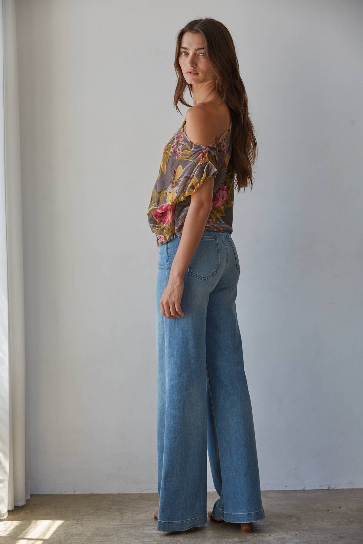 A woman stands in profile wearing a colorful floral cold-shoulder top and wide-leg blue jeans against a plain light gray wall.