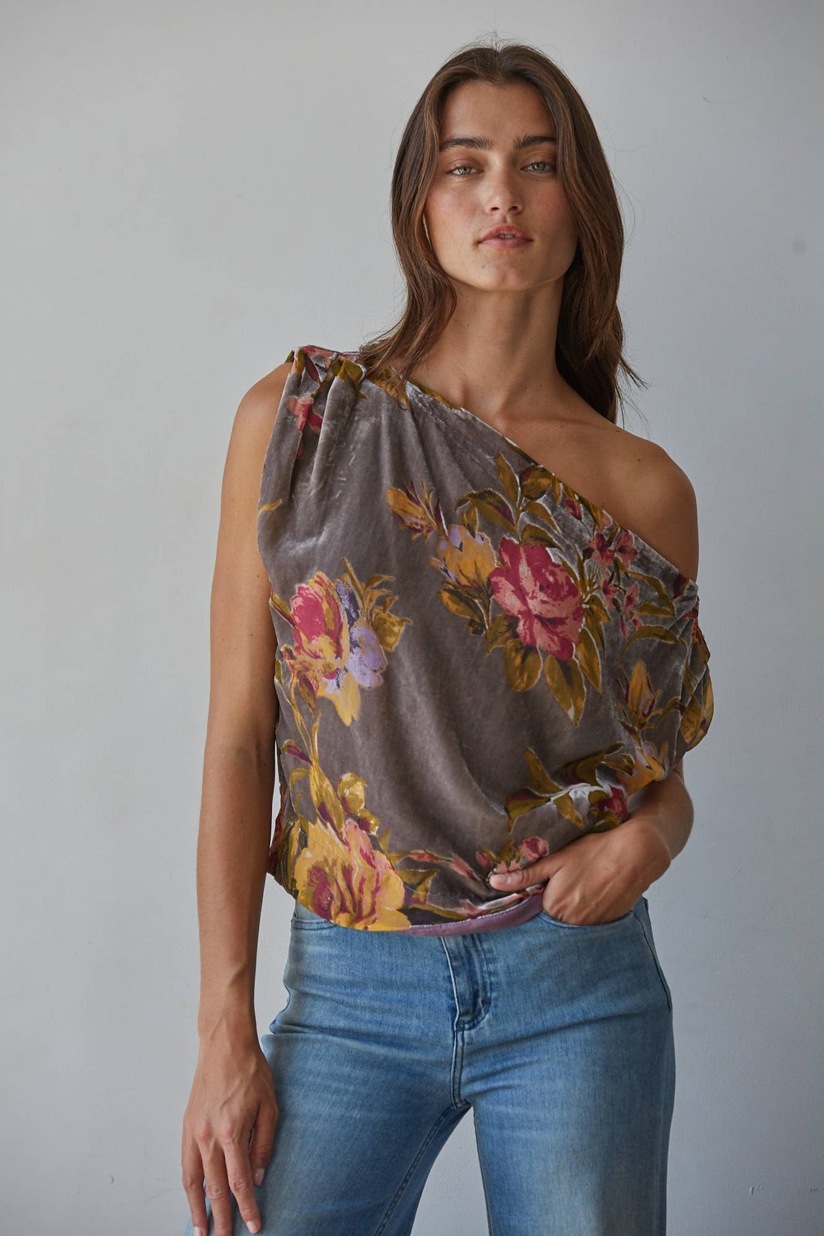 A woman wearing a draped, one-shoulder floral top in muted brown, pink, and yellow tones, paired with blue jeans, standing against a plain light gray wall.