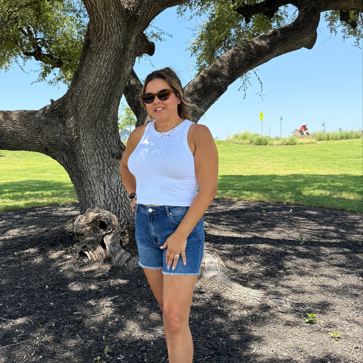 A woman posing outdoors under a large tree, wearing a white sleeveless top, blue denim shorts, and white sneakers.