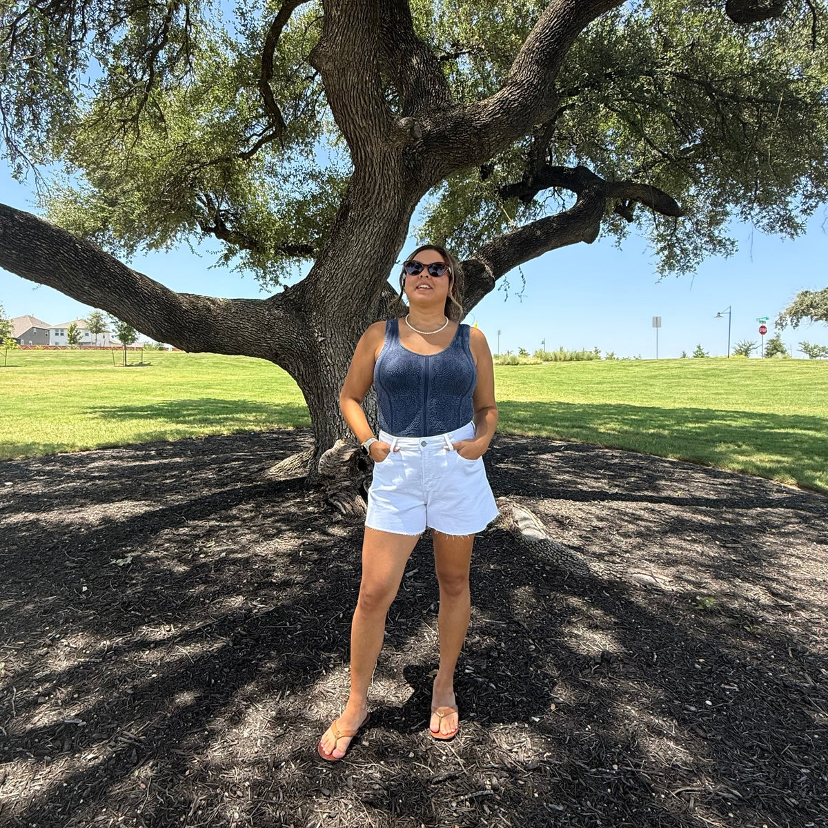 A woman stands under a large tree in a park, wearing a navy blue tank top, white shorts, and sunglasses. The background shows a grassy field and blue sky.