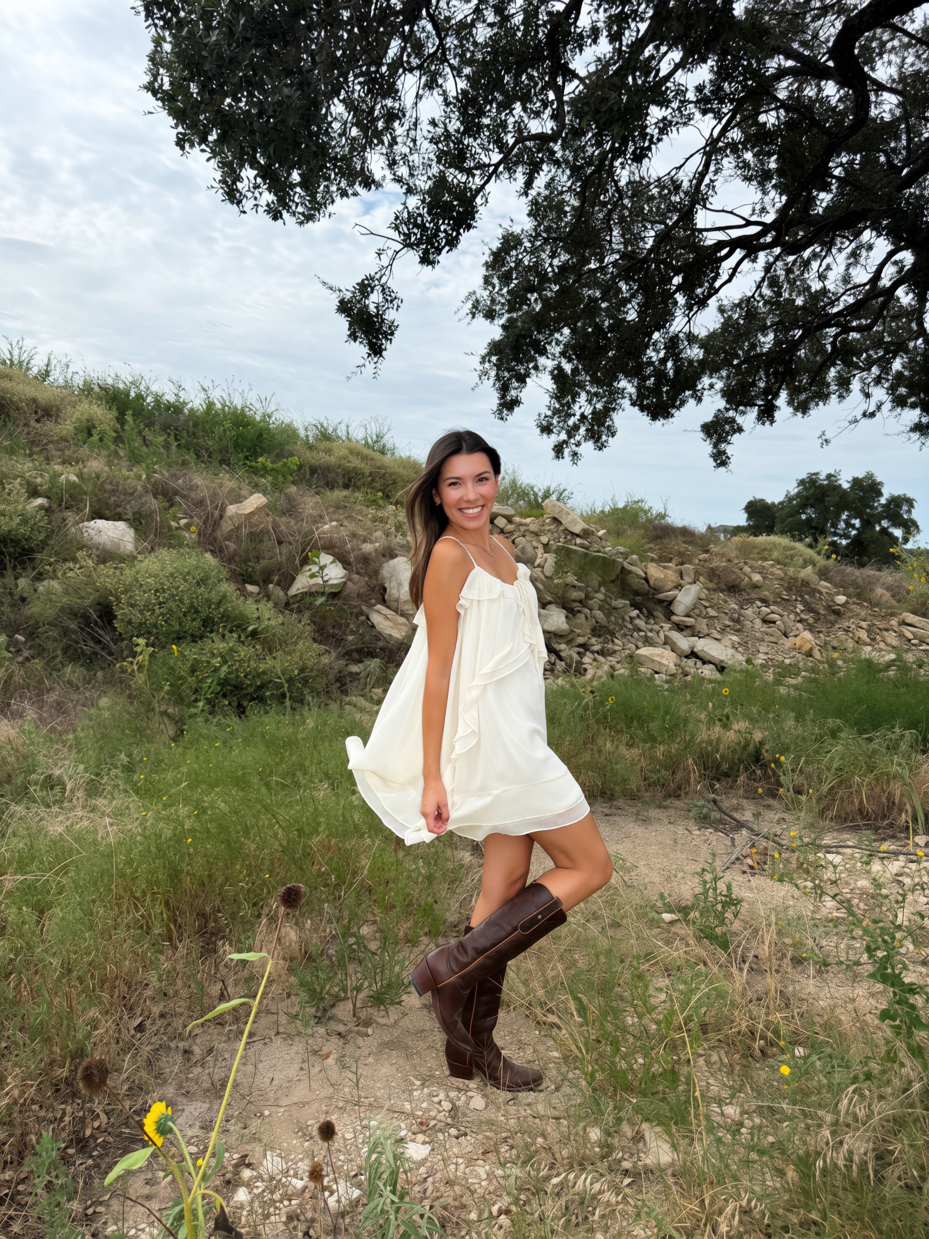 A woman wearing a white ruffle western Texas rodeo sleeveless mini dress and brown cowboy boots stands outdoors on a grassy hillside under a tree.