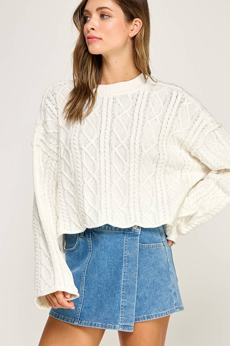 A woman wearing a cream cable-knit cropped sweater with long bell sleeves and a blue denim mini skirt, photographed against a light gray studio background.