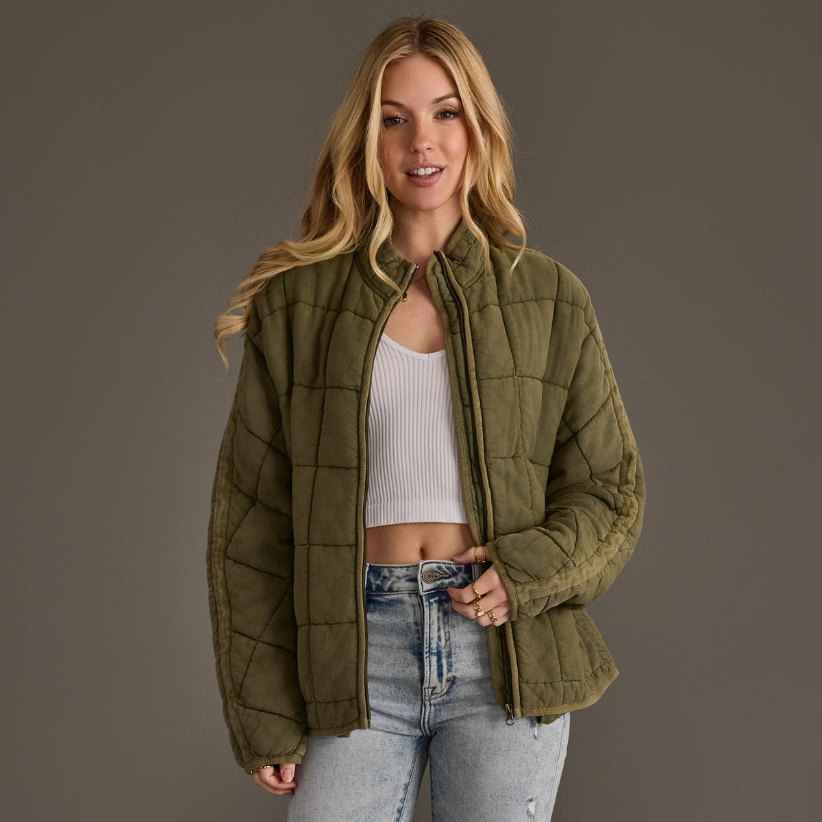 A woman modeling a quilted olive green jacket over a white ribbed crop top and light wash blue jeans, set against a solid dark gray studio background.