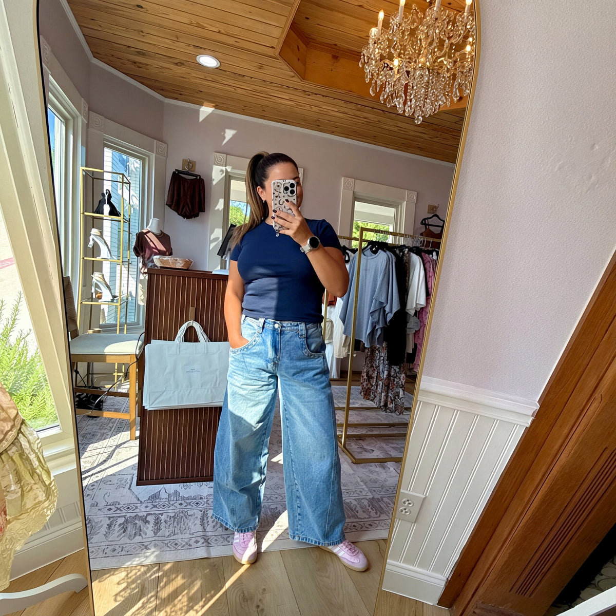 A woman takes a mirror selfie in a boutique showing a pair of wide-leg light blue denim jeans, a navy blue t-shirt, and pink sneakers. The boutique has light wood floors, a wooden ceiling with a crystal chandelier, and clothing racks in the background.