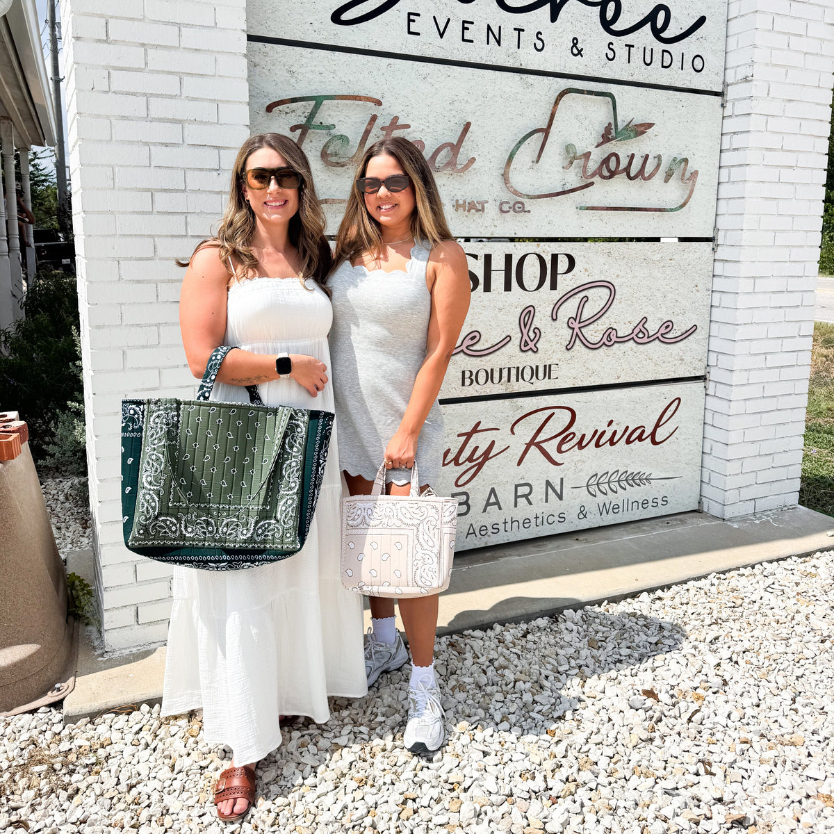 Two women posing in front of a white brick sign that reads 'Soirée Events & Studio' with additional storefront text behind them. The woman on the left wears a white sleeveless dress and holds a dark green patterned tote bag, while the woman on the right wears a light gray dress and holds a white patterned tote bag.