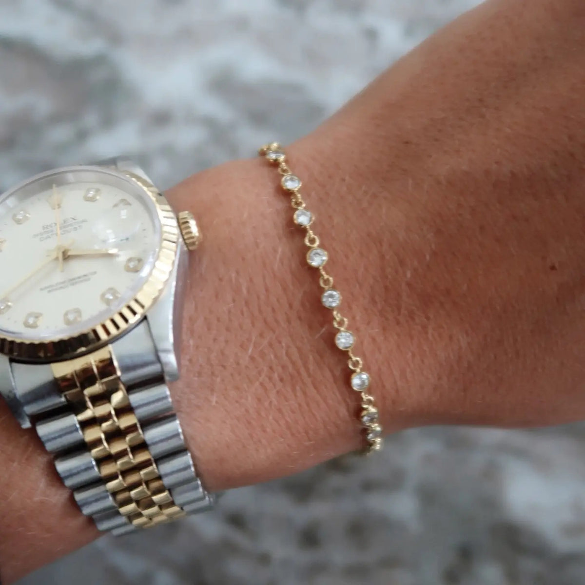 A gold chain bracelet with clear round stones worn on a person's wrist next to a two-tone stainless steel and gold watch.