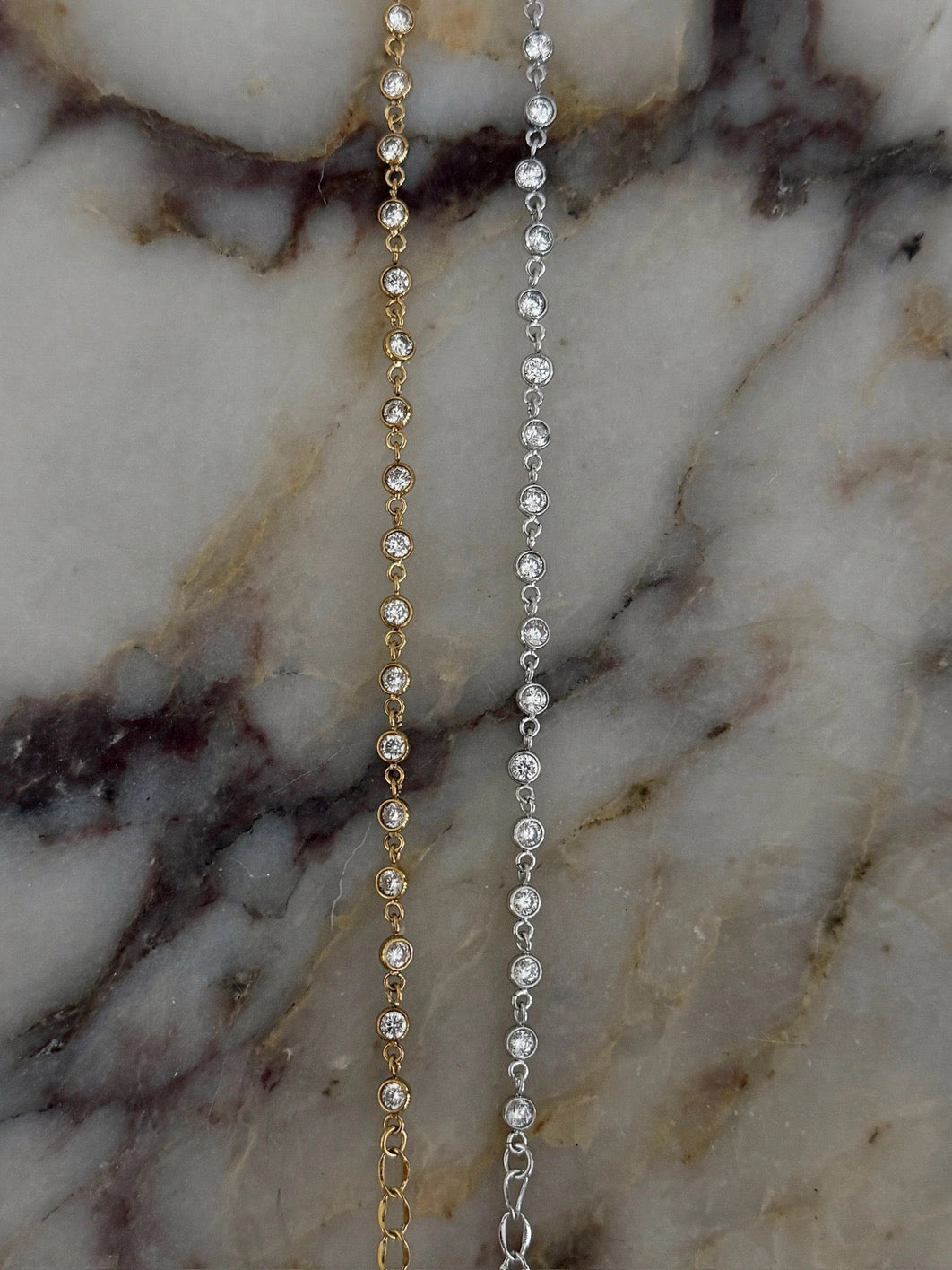 Two delicate chain bracelets, one gold-toned and one silver-toned, laid flat on a white and gray marble surface.