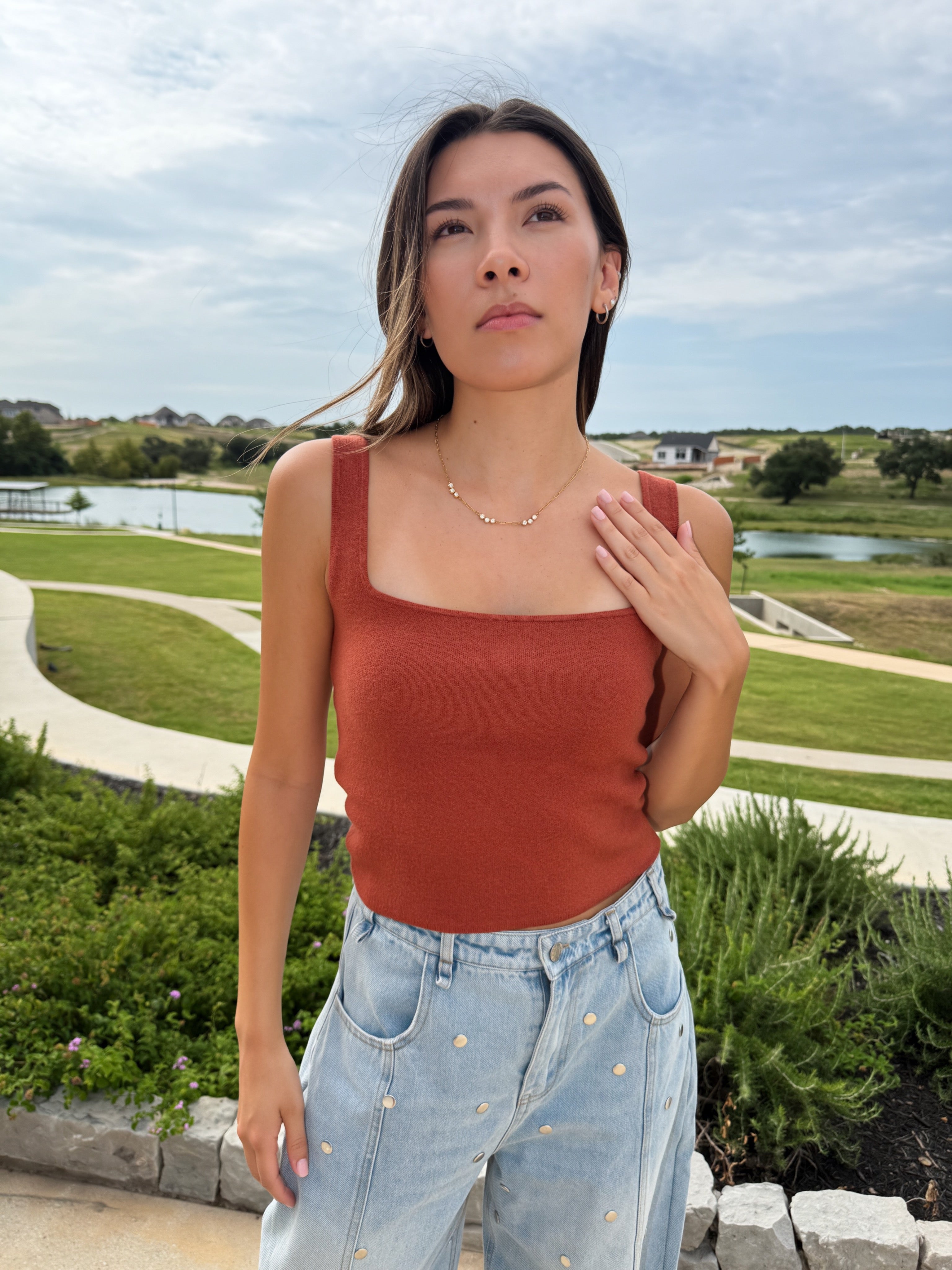 A woman wearing a burnt orange Texas longhorn game day ribbed knit tank top and light wash denim jeans with silver studs, standing outdoors in front of a landscaped park with a lake and green grass.