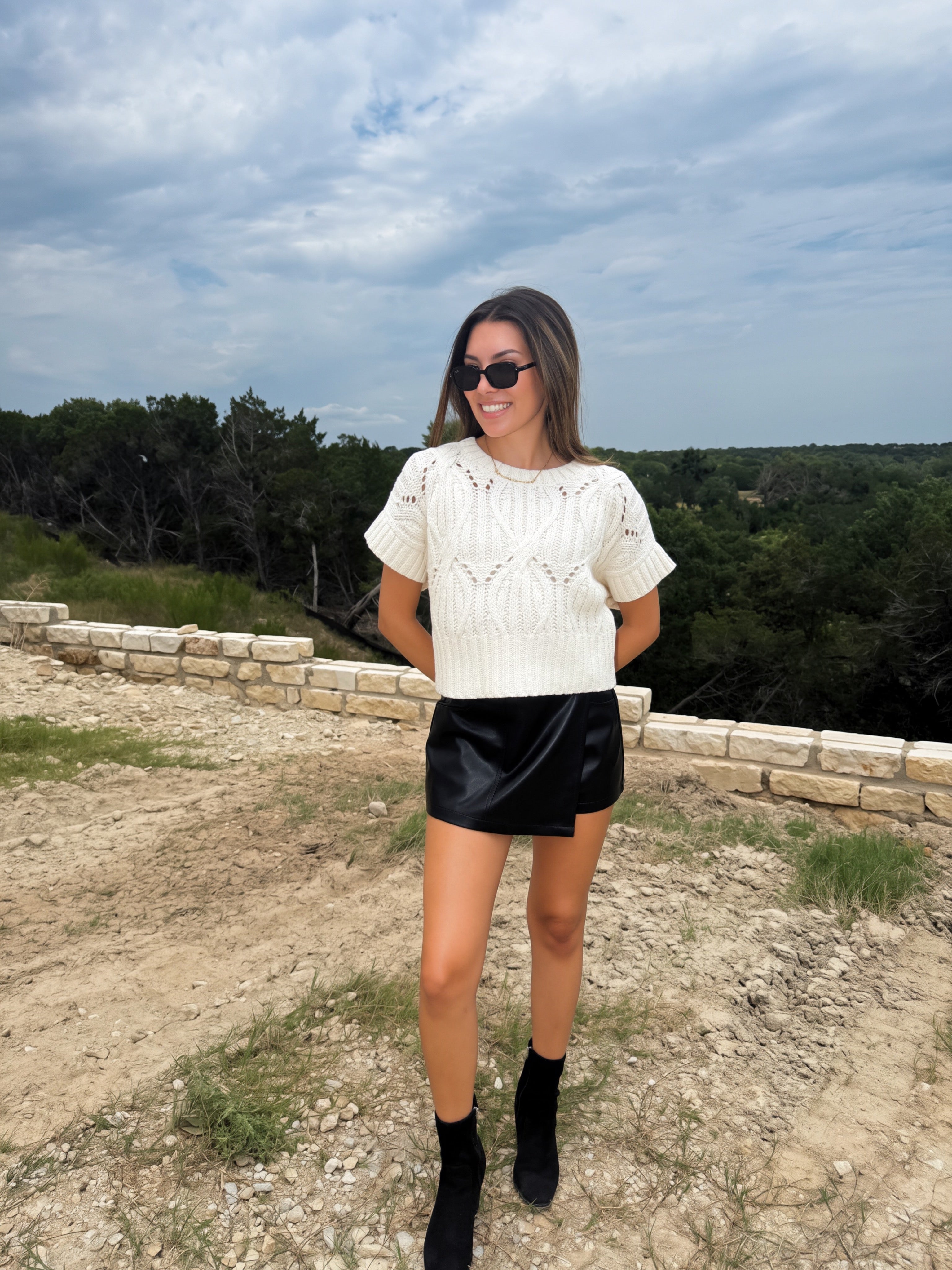 A woman wearing a cream cable-knit short-sleeve top, a black faux-leather mini skirt, and black ankle boots, standing outdoors on a dirt path with a stone wall and trees in the background.