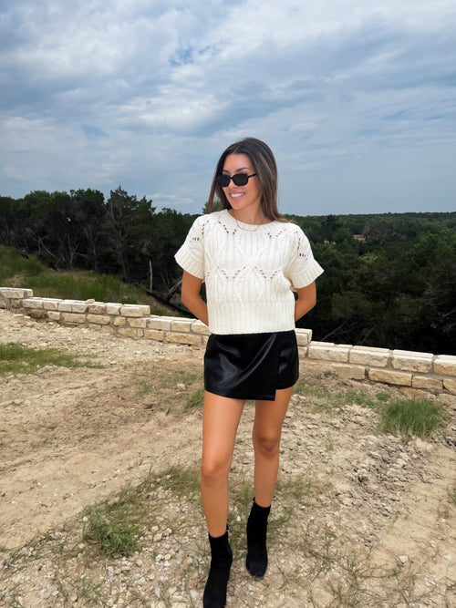 A woman wearing a cream cable-knit short-sleeve top, a black faux-leather mini skirt, and black ankle boots, standing outdoors on a dirt path with a stone wall and trees in the background.