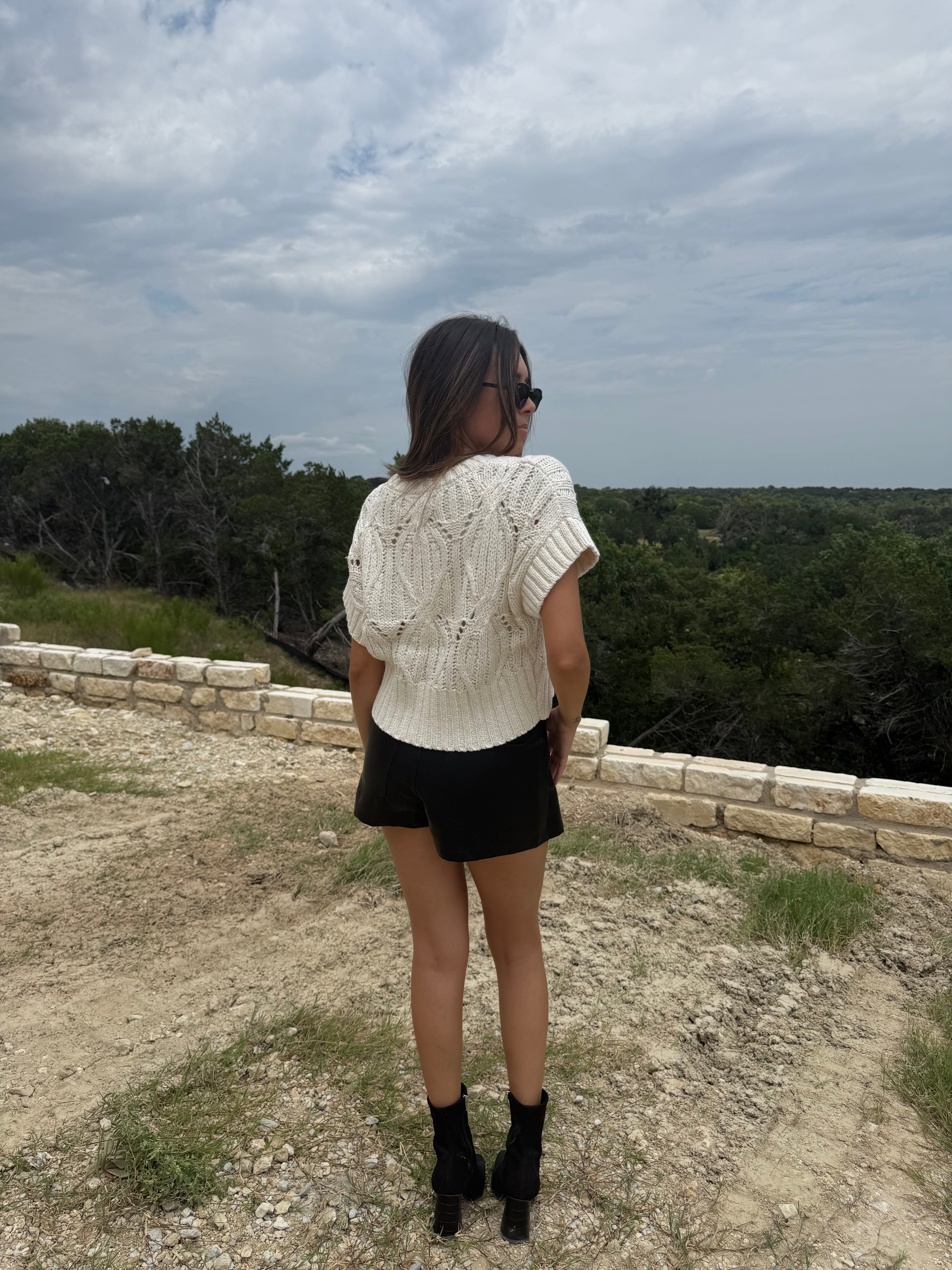A woman is seen from behind wearing a cream-colored textured knit short-sleeve top, black shorts, and black ankle boots, standing outdoors on a dirt path with a stone wall and trees in the background under a cloudy sky.