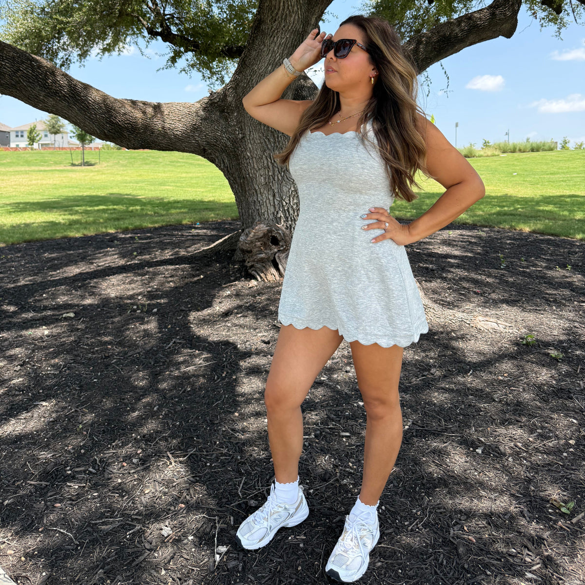 A woman posing outdoors under a large tree wearing a light grey mini dress, white sneakers, and white socks.