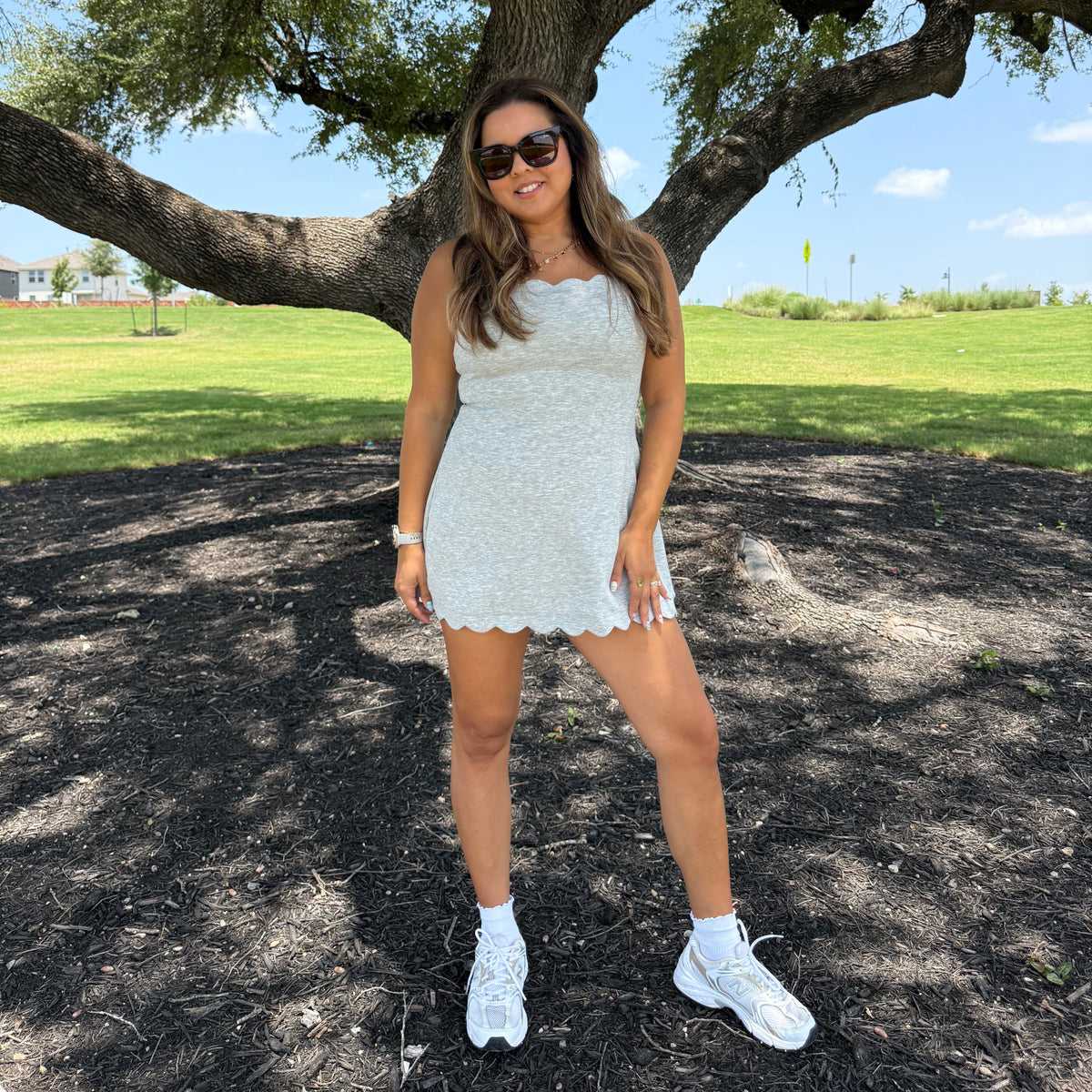 A woman posing outdoors under a large tree, wearing a grey sleeveless dress, white sneakers, and sunglasses on a grassy park lawn.