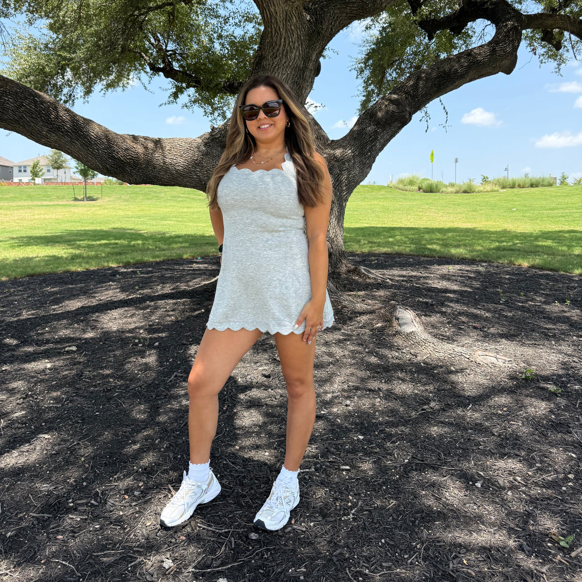 A woman wearing a scalloped grey athletic mini dress, white sneakers, and sunglasses stands under a large tree in a park with green grass and blue sky in the background.