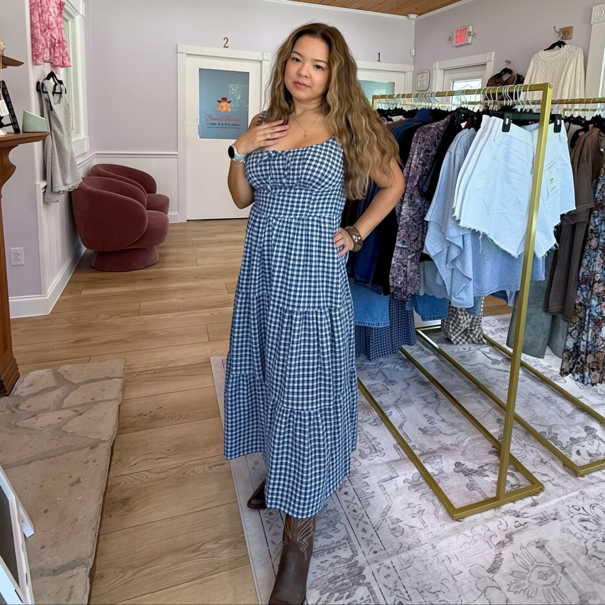 A woman modeling a blue and white gingham sleevely midi dress inside a boutique clothing store with wooden ceiling, clothing racks, and a crystal chandelier.