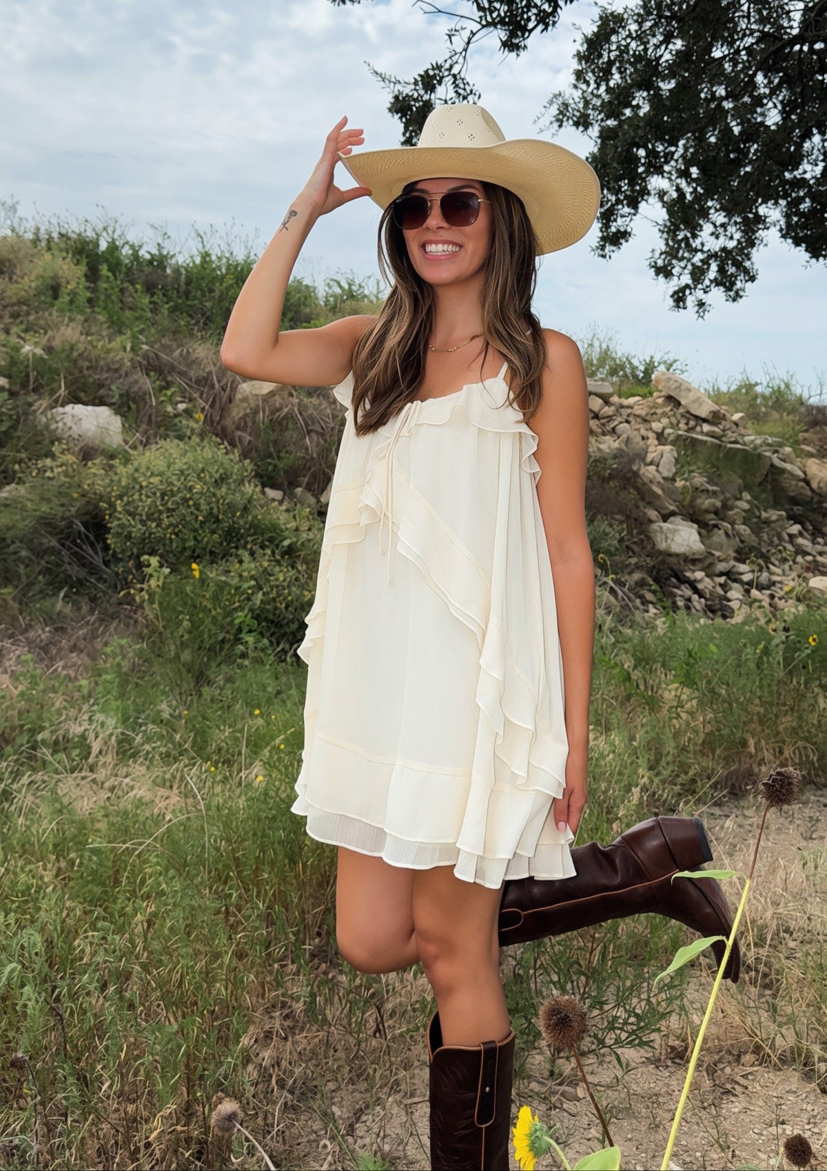 A woman wearing a cream ruffled sleeveless western style fall dress, a wide brim straw cowboy hat, and brown cowgirl boots stands outdoors in a grassy field under a cloudy sky.