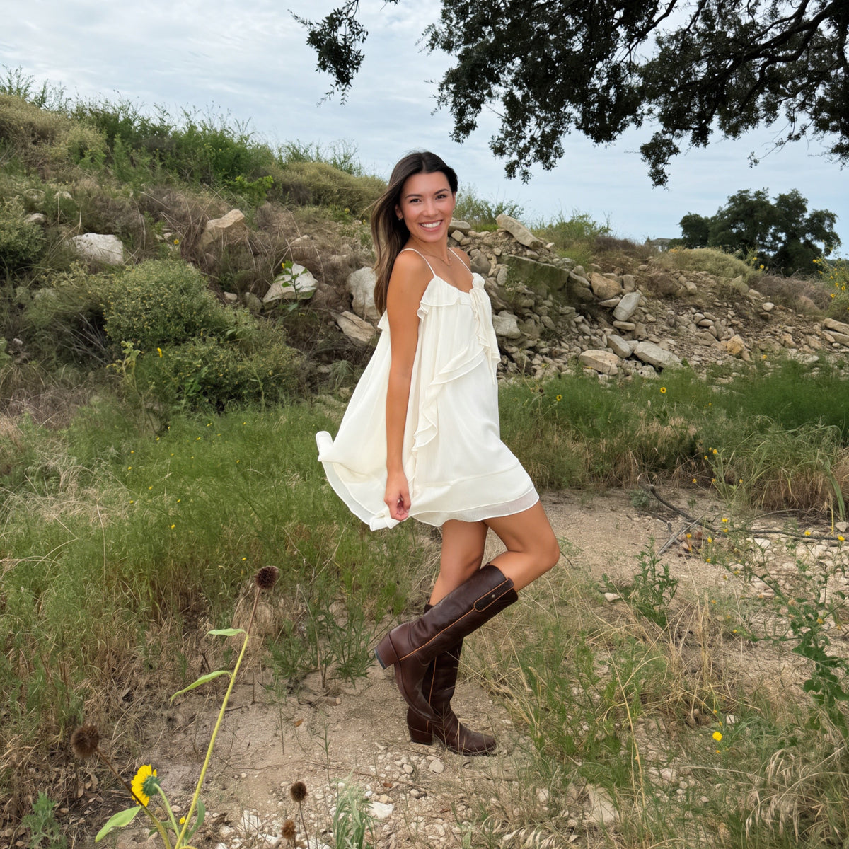 A woman wearing a white ruffle western Texas rodeo sleeveless mini dress and brown cowboy boots stands outdoors on a grassy hillside under a tree.