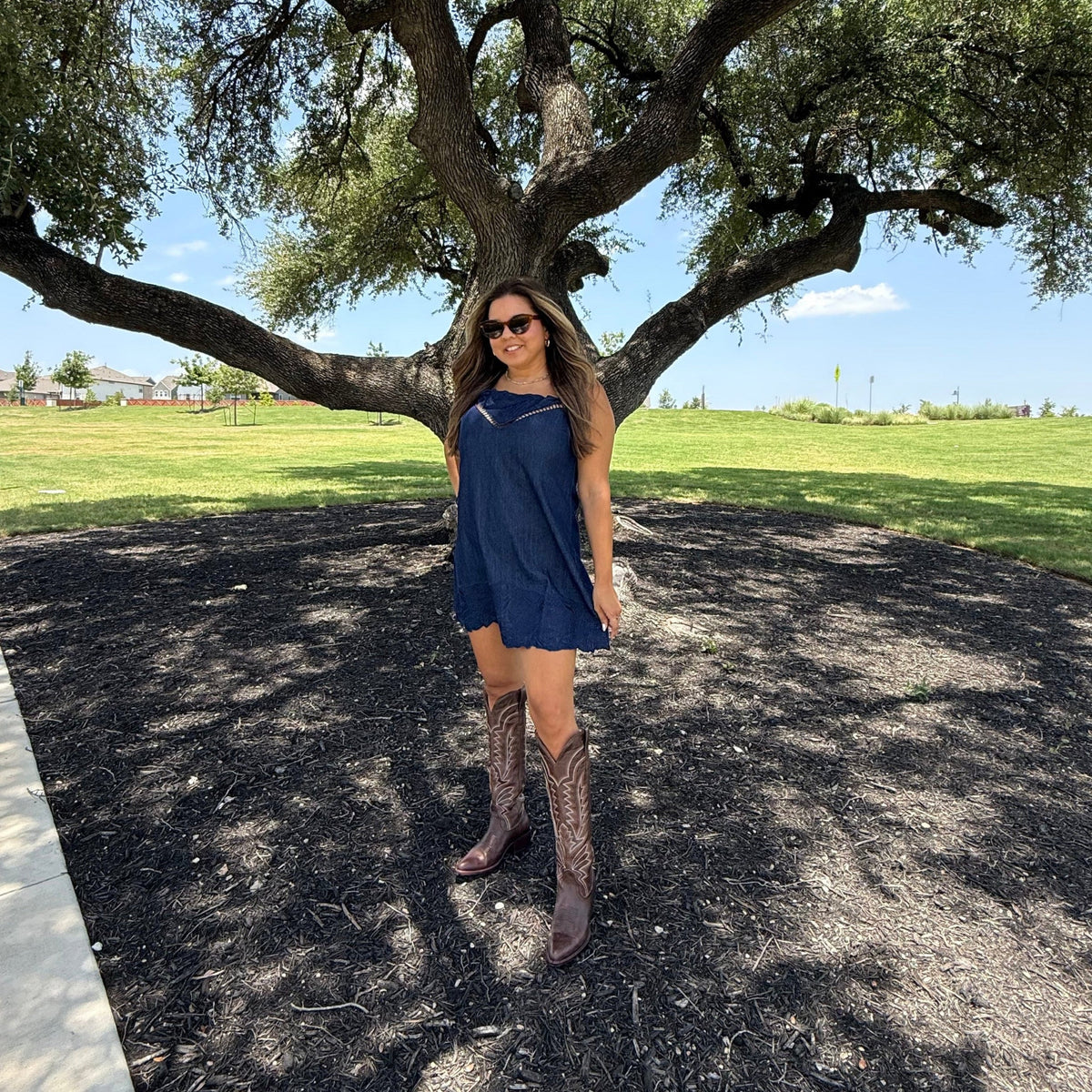 A woman wearing a navy blue sleeveless dress and brown cowboy boots stands outdoors under a large tree on a paved path with grass in the background.
