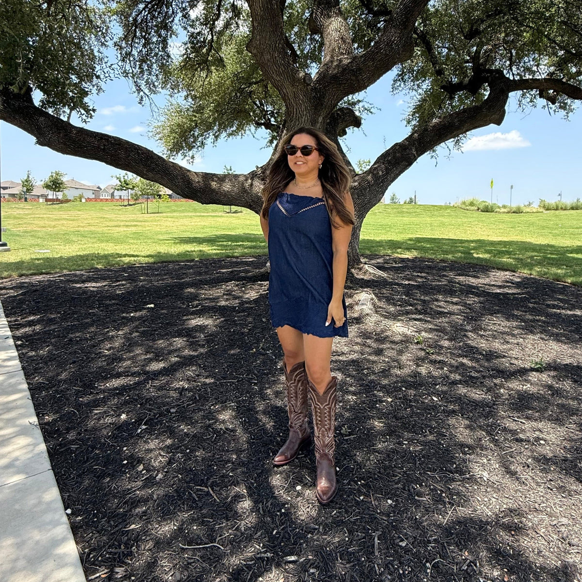 A woman posing outdoors under a large tree, wearing a navy blue sleeveless dress and brown lace-up boots on a paved path.