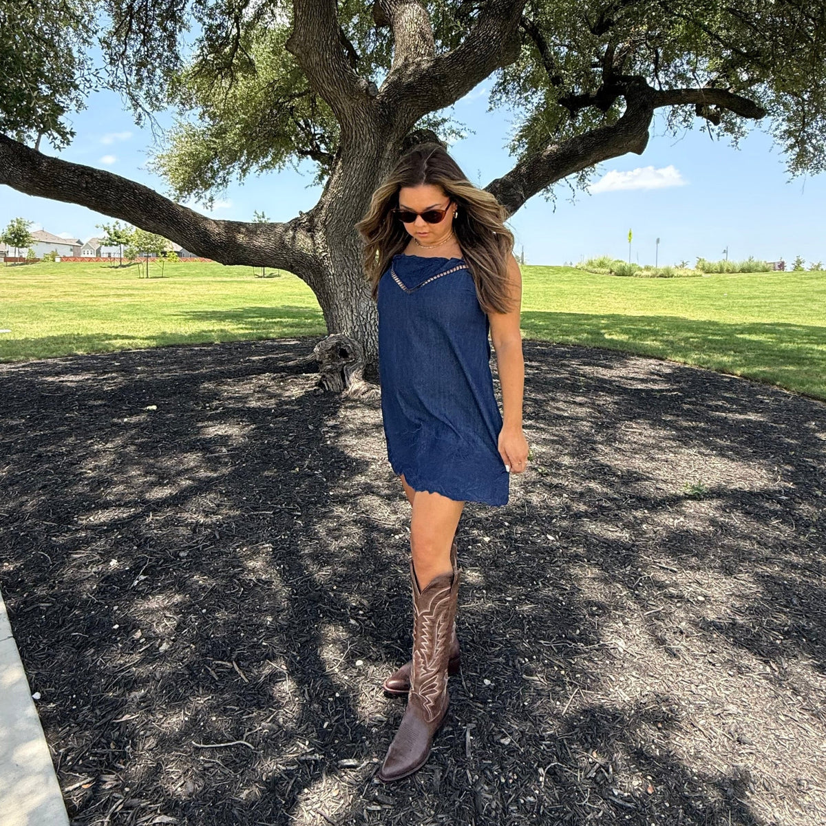 A woman wearing a sleeveless navy blue dress and brown cowboy boots stands outdoors under a large tree on a paved path with grass in the background.