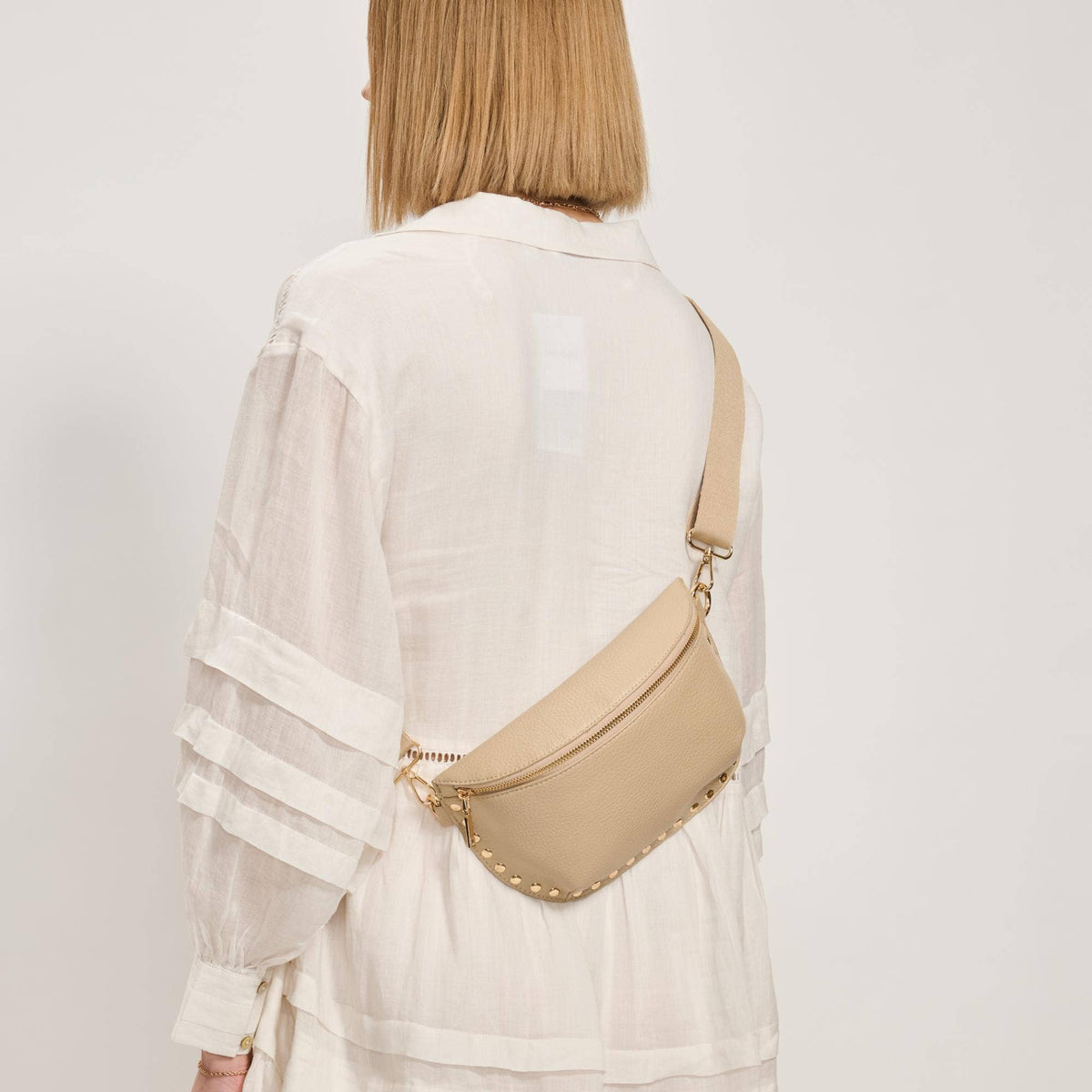 Person wearing a beige crossbody bag with studded details on a plain background