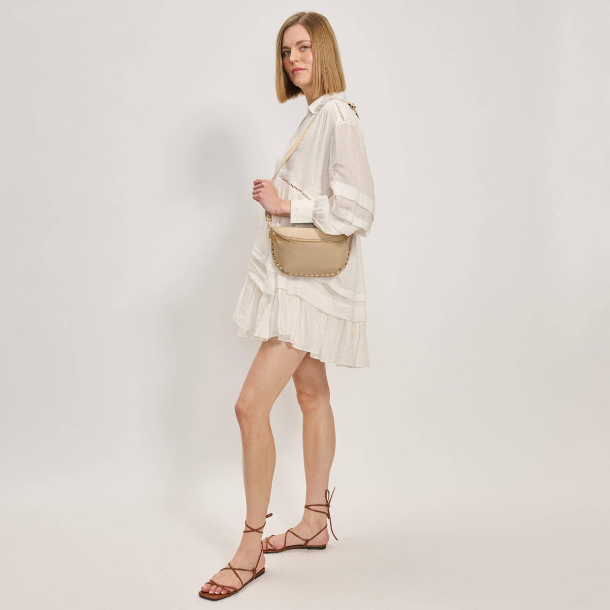 Woman wearing a white dress and beige sandals, holding a beige handbag on a plain background