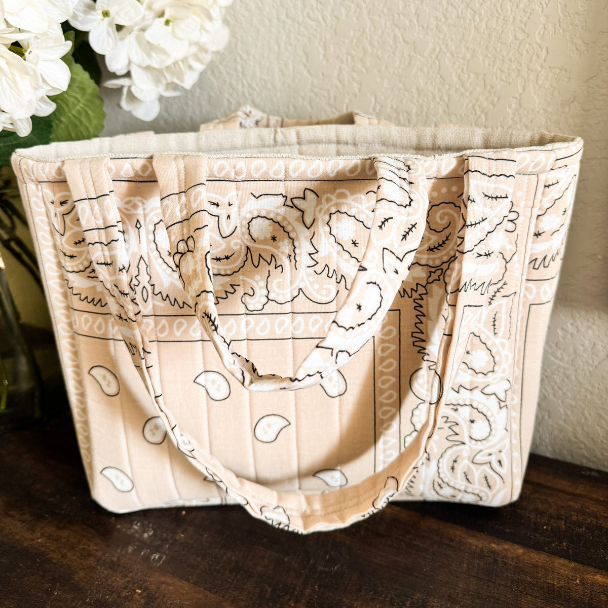 A beige fabric tote bag with black line-art illustrations of a dog and paisley patterns, resting on a dark wooden surface against a textured beige wall with white flowers in the background.