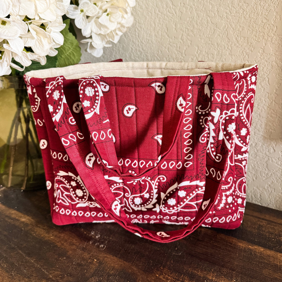 A red bandana print tote bag with white paisley patterns, resting on a dark wooden table against a beige wall with white flowers in the background.