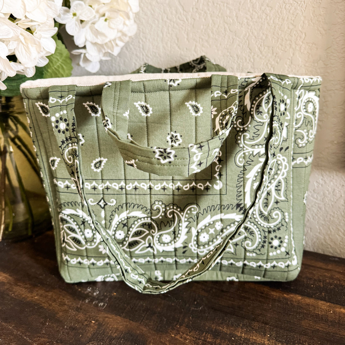 An olive green fabric tote bag with a white paisley bandana print, resting on a dark wooden table next to a vase of white flowers against a textured beige wall.