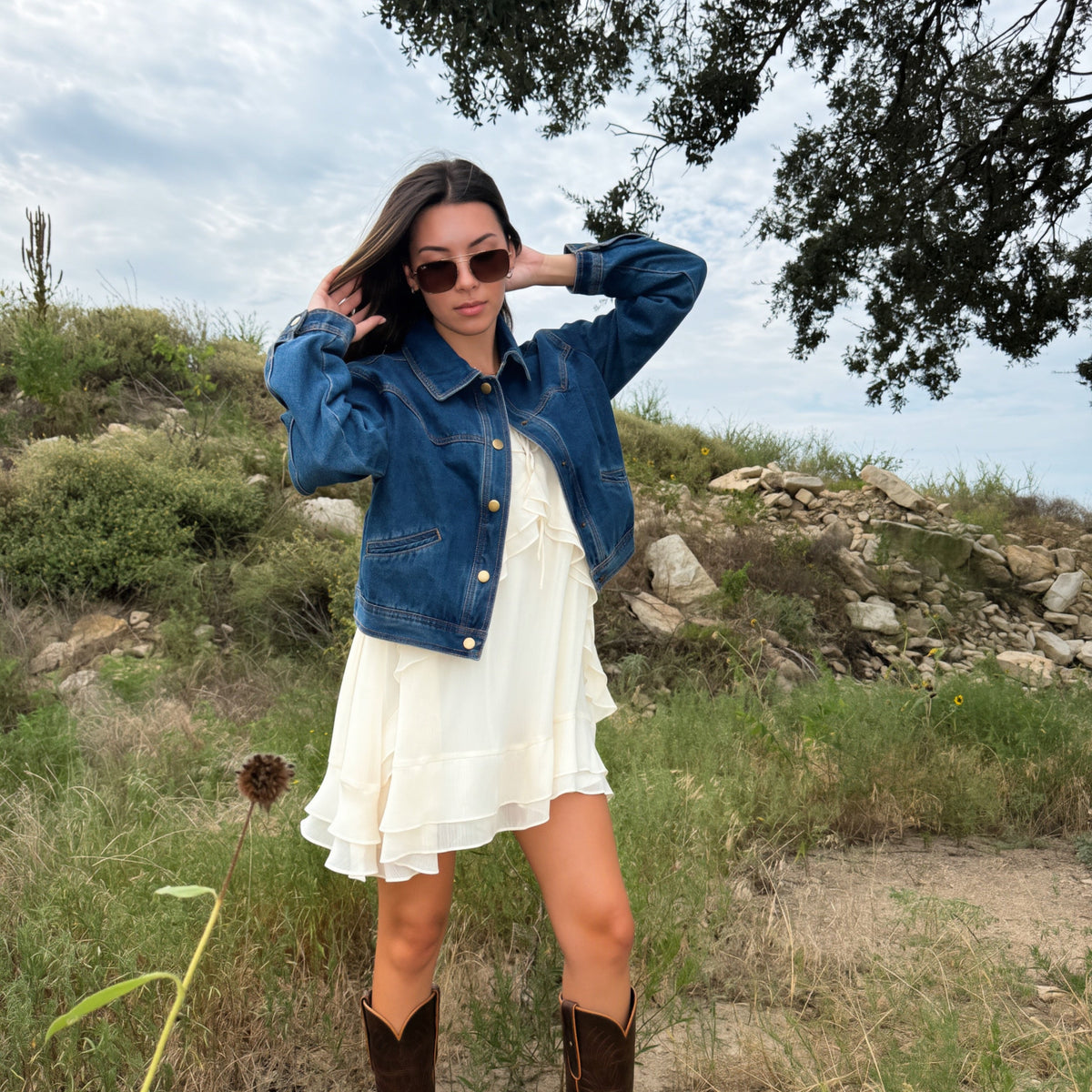 A woman stands outdoors on a grassy hill wearing a blue fall western style denim jacket over a white ruffled dress, with brown cowboy boots and sunglasses.
