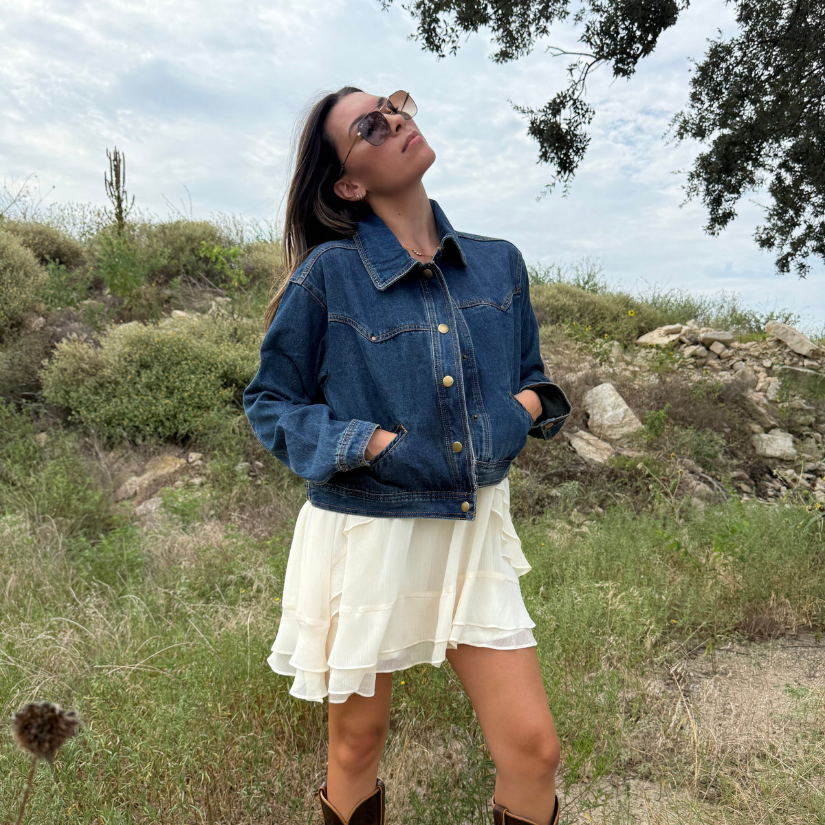 A woman wearing a dark blue denim jacket, cream ruffled mini dress, and brown cowboy boots stands outdoors in a grassy, rocky landscape under a cloudy sky. Fall Style Texas 