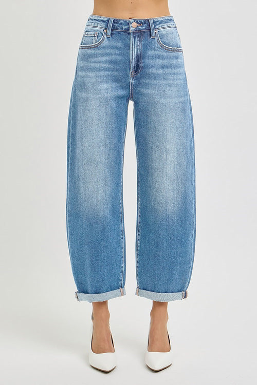 A model is wearing a pair of wide-leg, cropped blue denim jeans with rolled cuffs, paired with white pointed-toe heels, shown against a plain light gray studio background.