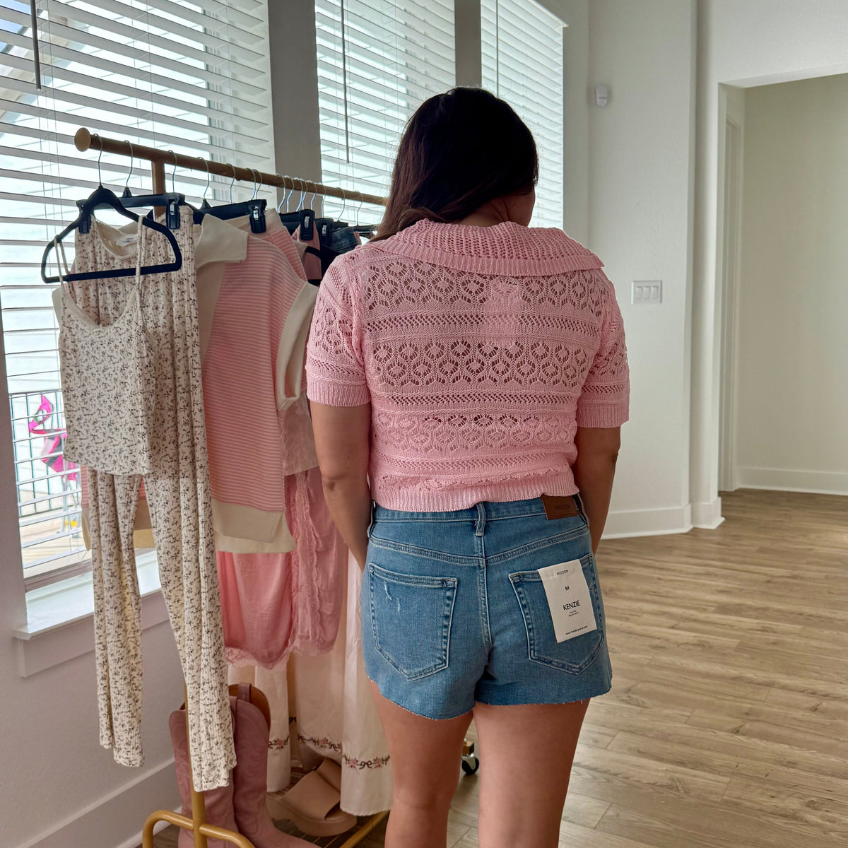 Woman in a pink sweater and denim shorts standing in front of a clothing rack.