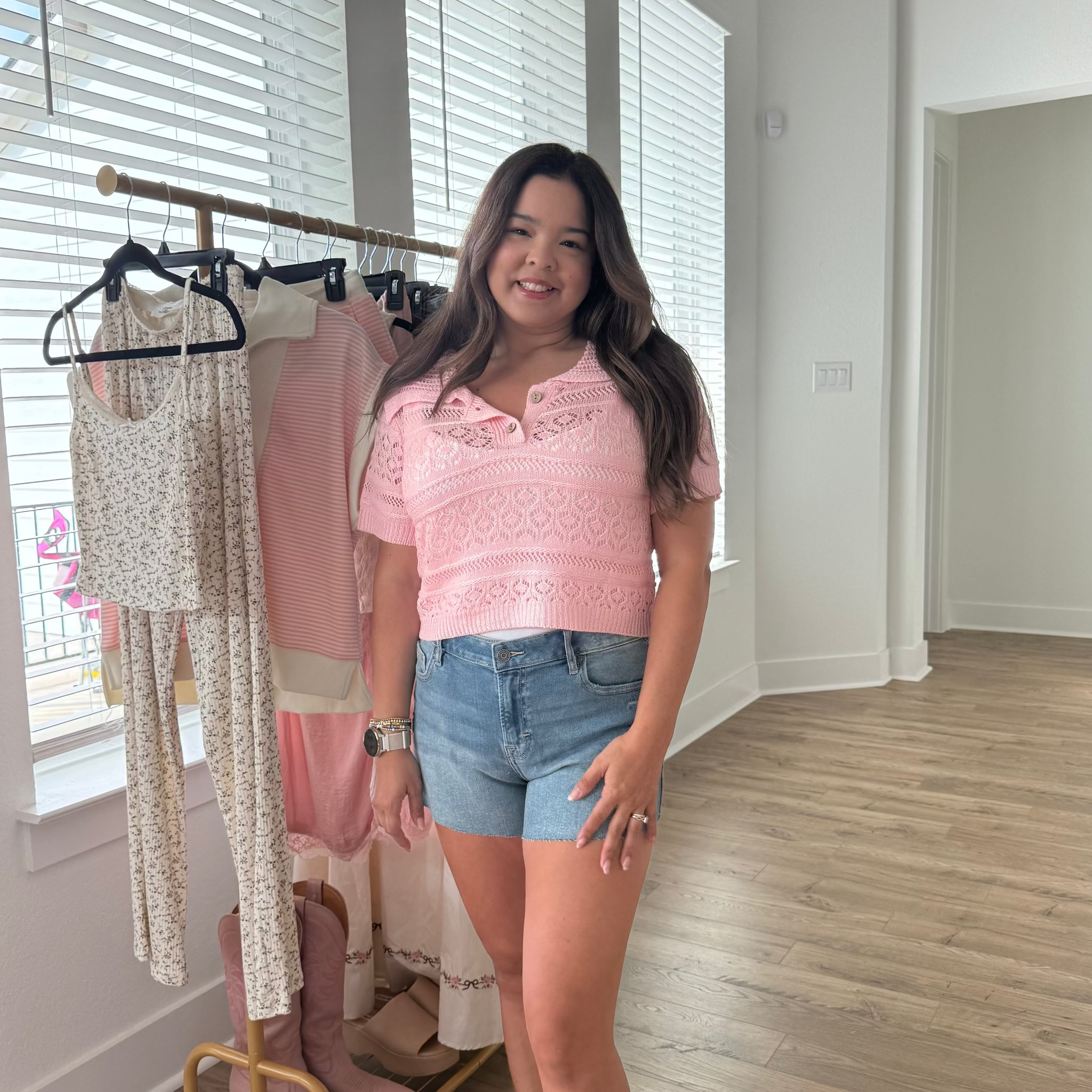 Woman in a pink top and denim shorts standing next to a rack of clothes in a room with wooden flooring.
