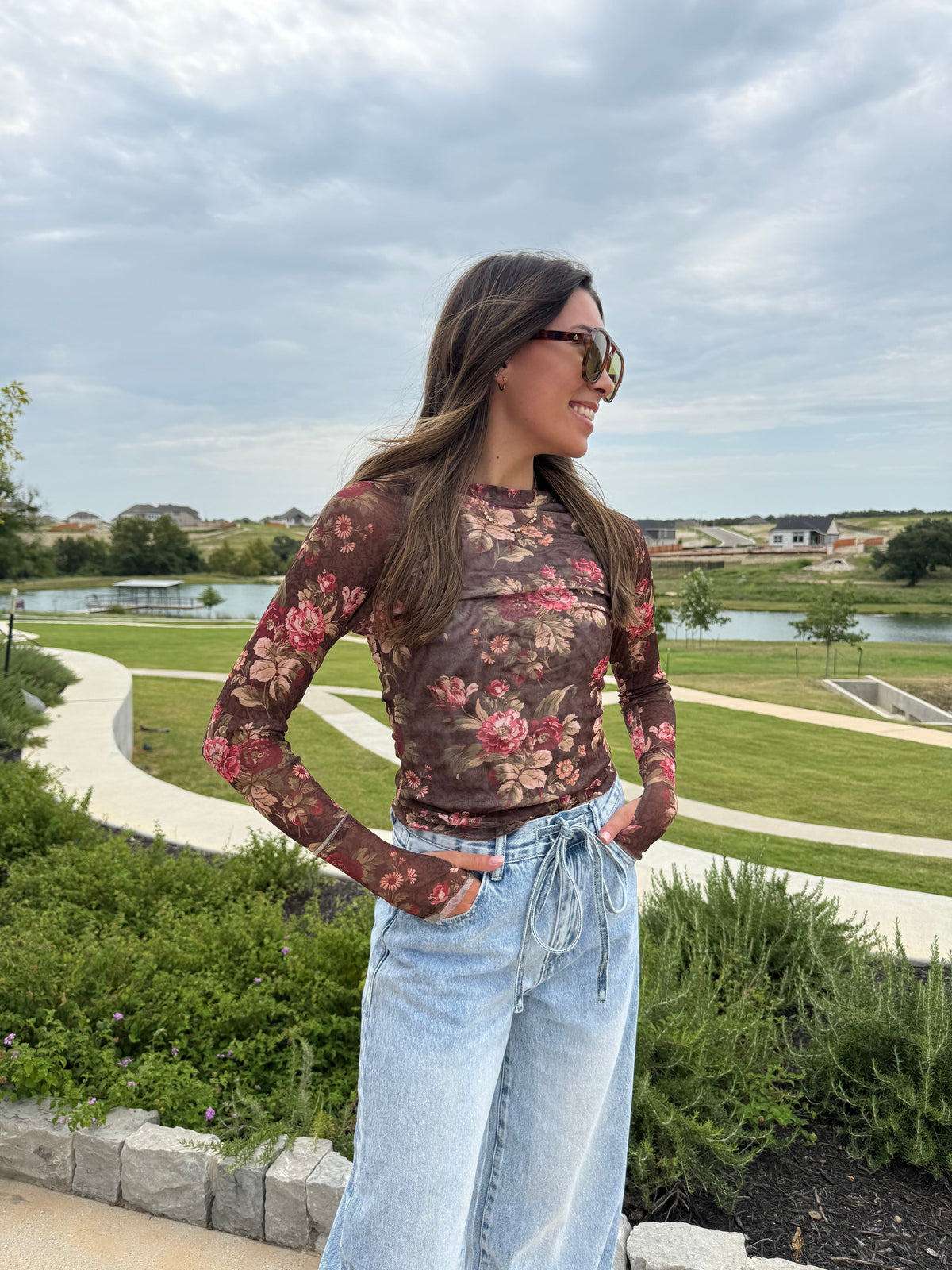 A woman wearing a brown floral long-sleeve mesh top and light blue wide-leg jeans stands outdoors in a park setting with a lake and green grass in the background.