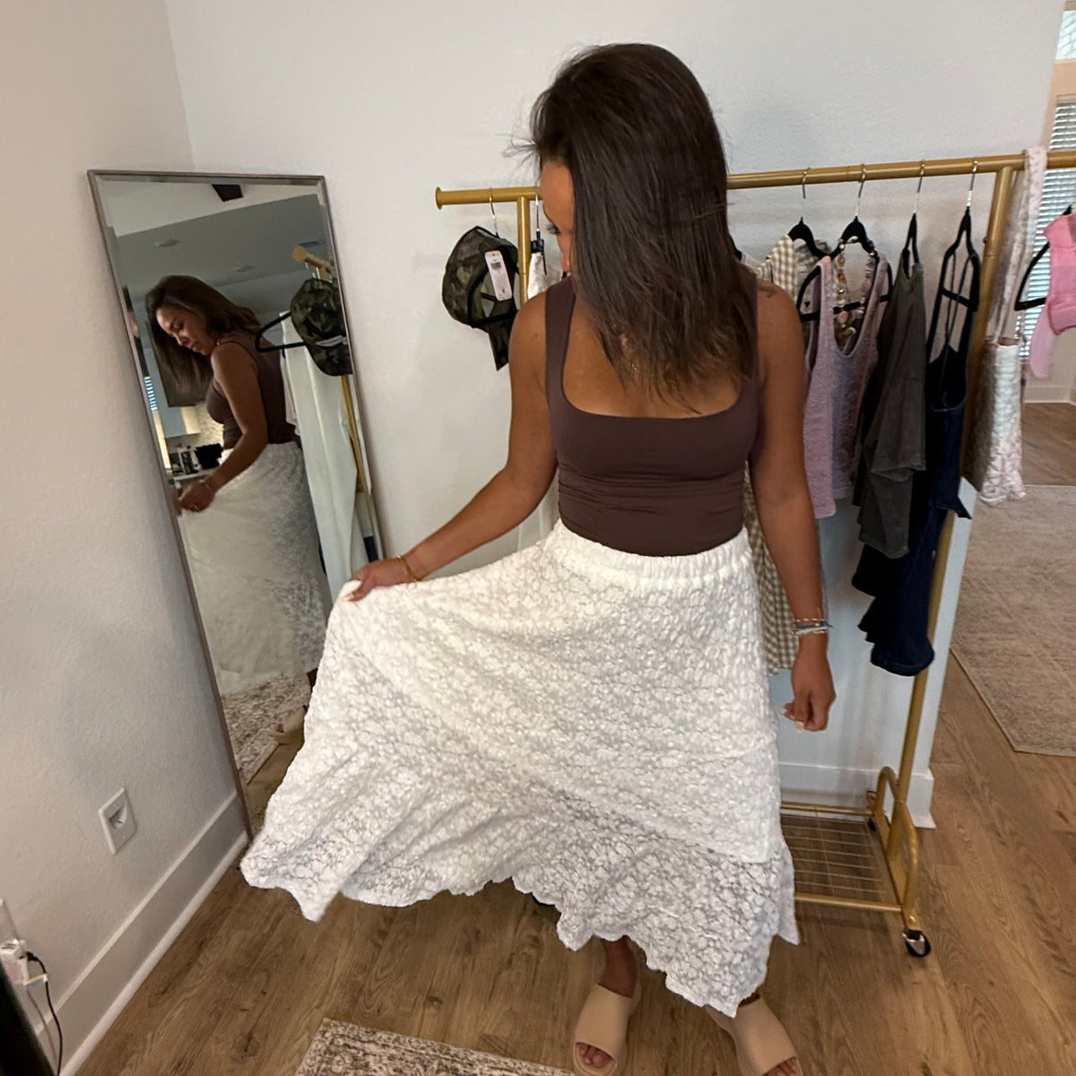 Woman in a brown top and white lace skirt standing in a room with a mirror and clothing rack.