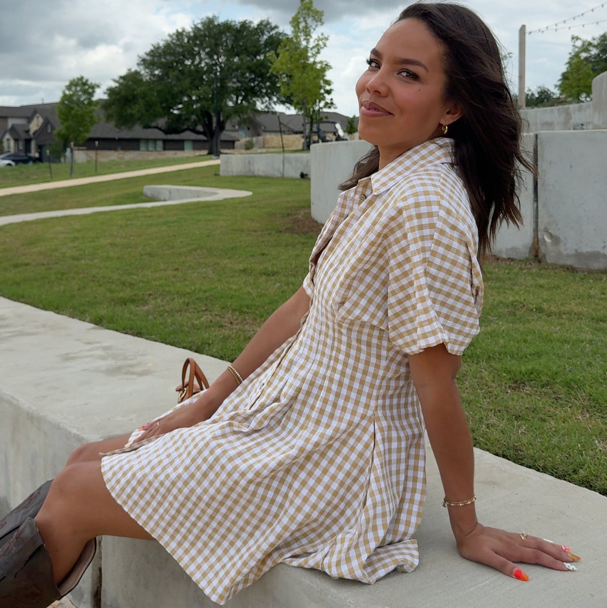 Woman in a checkered dress standing on a stone pathway with grass and trees in the background.