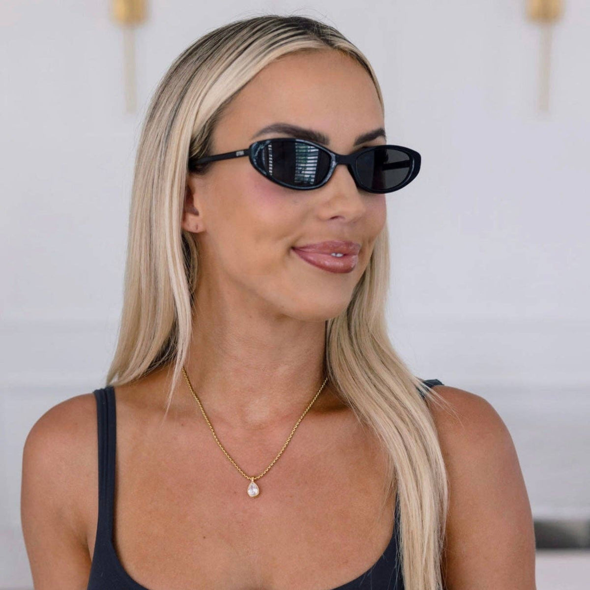 A blonde woman wearing black cat-eye sunglasses, a black tank top, and a delicate gold necklace with a small white bead pendant, photographed in a bright indoor setting.