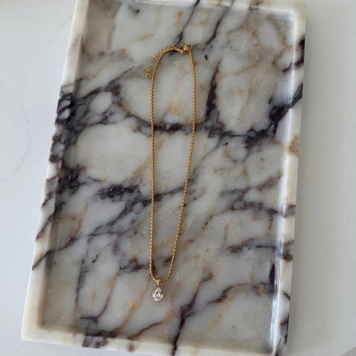 A delicate gold chain necklace with a small silver-toned teardrop-shaped pendant, displayed on a rectangular white and gray marble tray.
