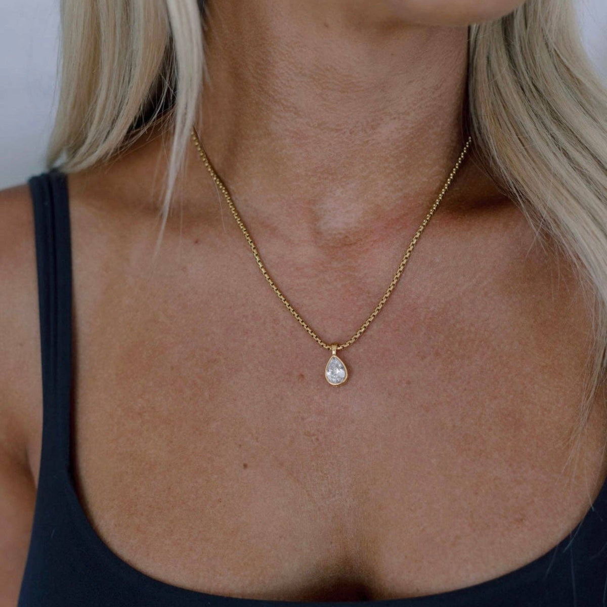 A close-up of a woman wearing a delicate gold chain necklace with a teardrop-shaped pendant, paired with a black tank top.