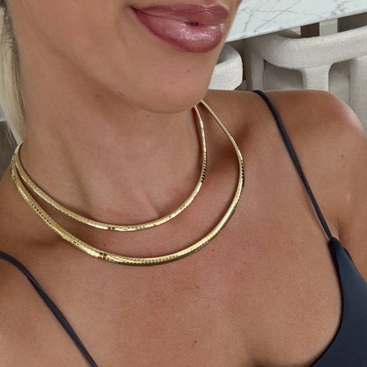 A woman wearing a dark navy blue spaghetti-strap camisole top and light-wash blue jeans, accessorized with a double-layered gold snake chain necklace and a silver bracelet.