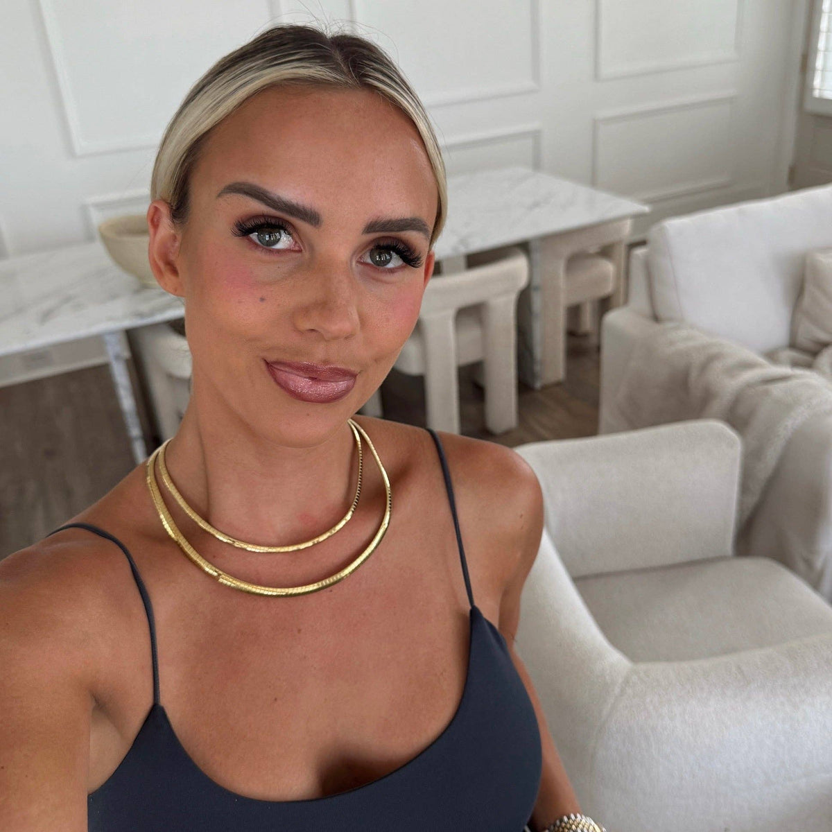 A woman wearing a navy blue camisole top and a double-layered gold chain necklace, posing in a bright, modern living room with white armchairs and a marble dining table in the background.