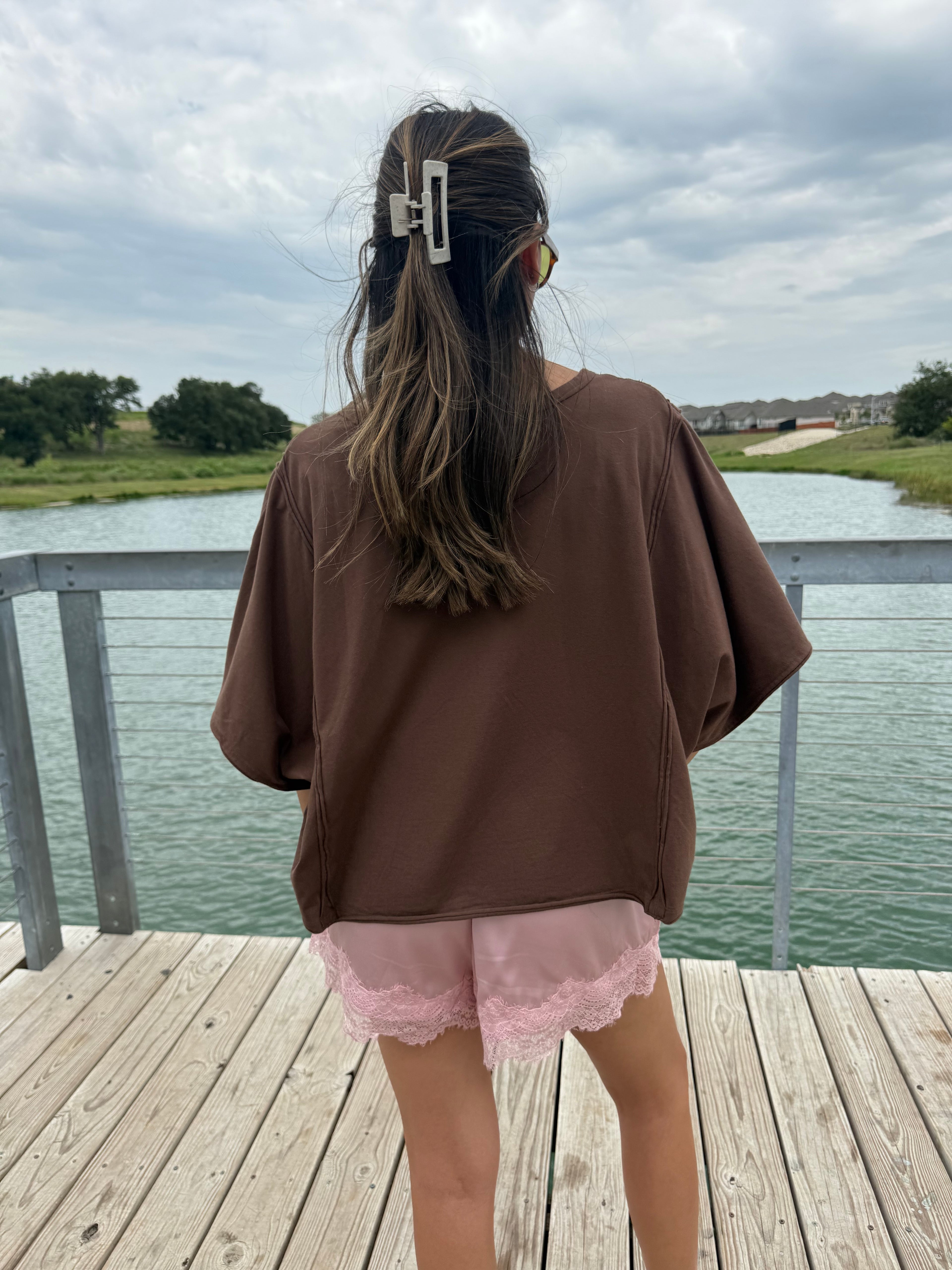 A person standing on a wooden deck overlooking a lake, wearing an oversized brown short-sleeve top and light pink lace-trim shorts, with a white hair clip holding their hair up.