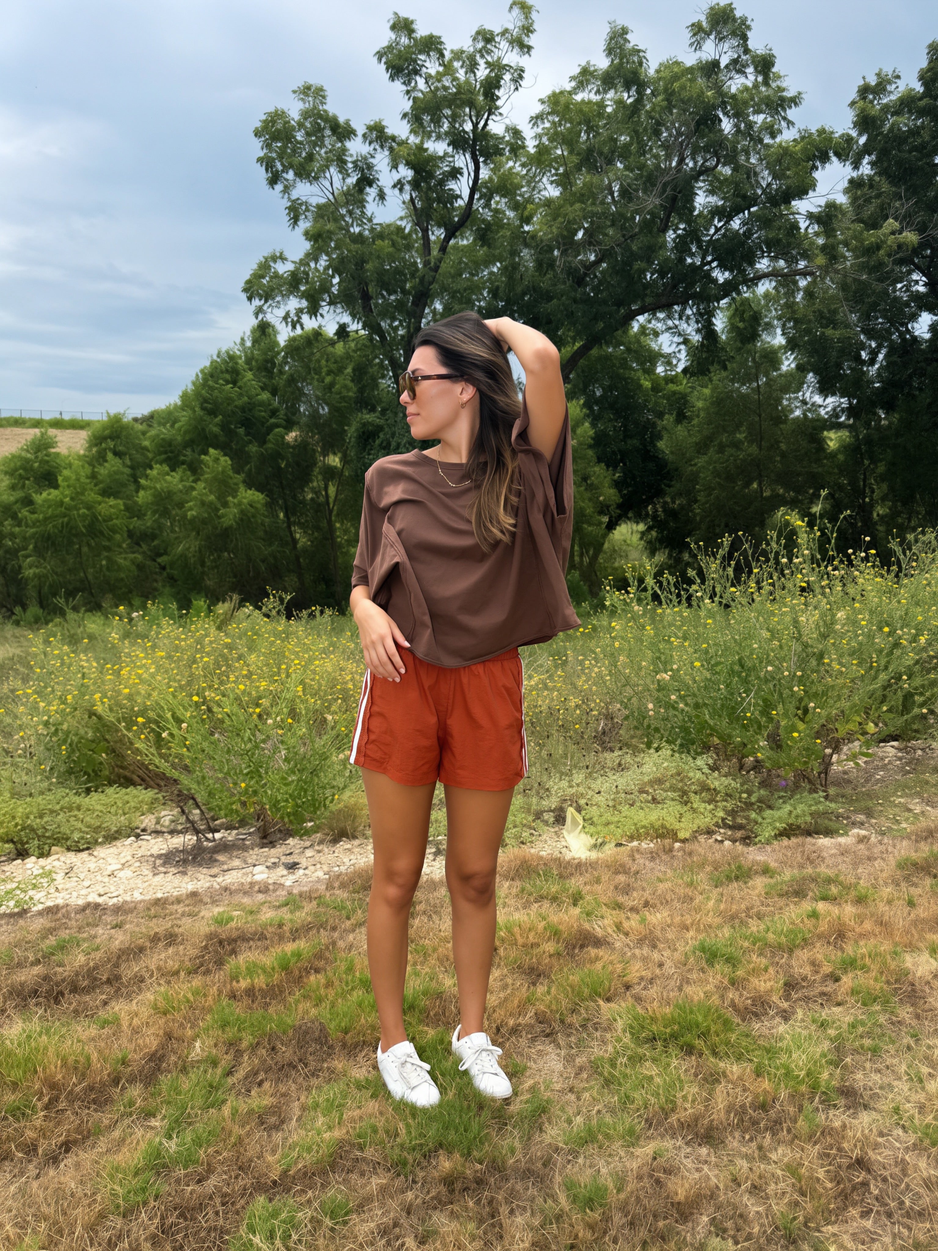 A woman stands outdoors in a grassy field with trees and shrubs in the background, wearing a loose brown t-shirt, rust-orange athletic shorts, and white sneakers.